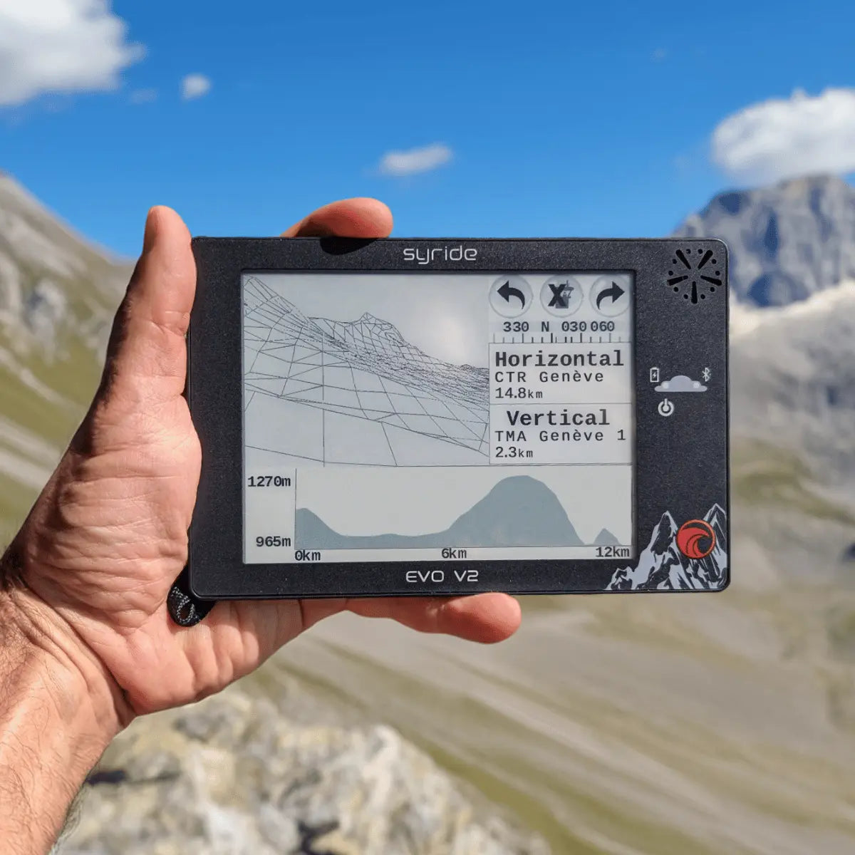 Hand hält das Syride Evo V2 Variometer für Gleitschirmflieger mit topografischer Karte und Fluginformationen vor Bergkulisse bei klarem Himmel.