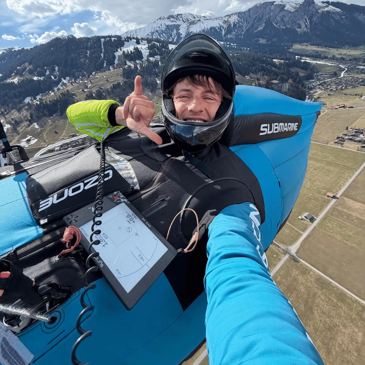 Piloten im Gleitschirm mit Helmunterstützung und Syride Evo V2 Variometer während eines Hike & Fly Fluges über Berglandschaft bei klarem Wetter.