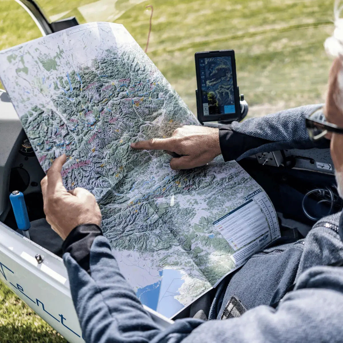 Mann im Segelflugzeug studiert eine detaillierte Karte des Alpenraums, mit einem Finger auf die Bergregion zeigend, daneben ein Tablet mit Navigations-App.
