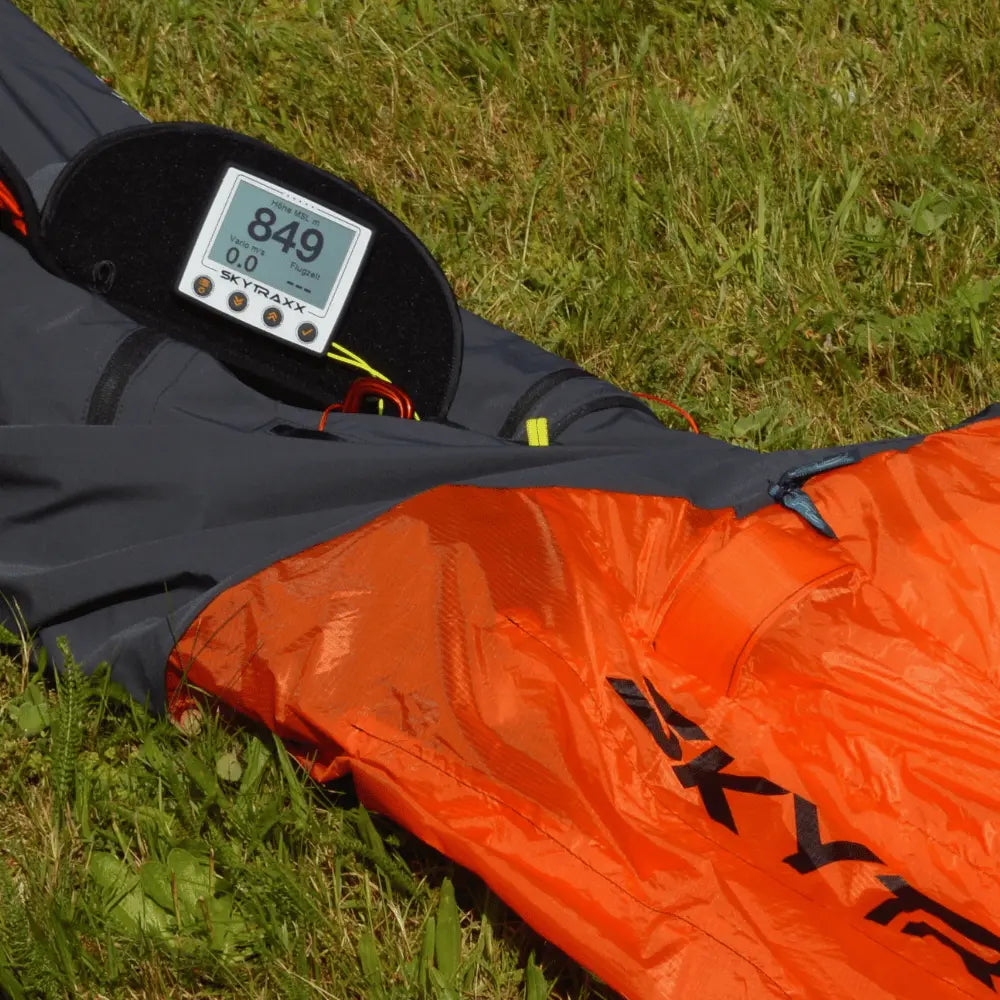 Skytraxx 5 mini - Vario in schaumstoffgepolstertem Etui auf orangener Gleitschirmtasche, ideal für Gleitschirmflüge mit FANET und FLARM, präsentiert auf grasigem Untergrund.
