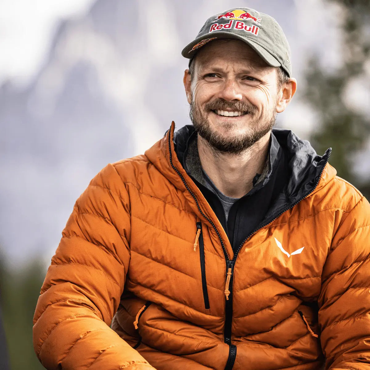 Porträt eines lächelnden Mannes mit roter Daunenjacke und Red Bull Cap in einer Berglandschaft, beworben für das Buch Wanderbird Strategie von Paul Guschlbauer.