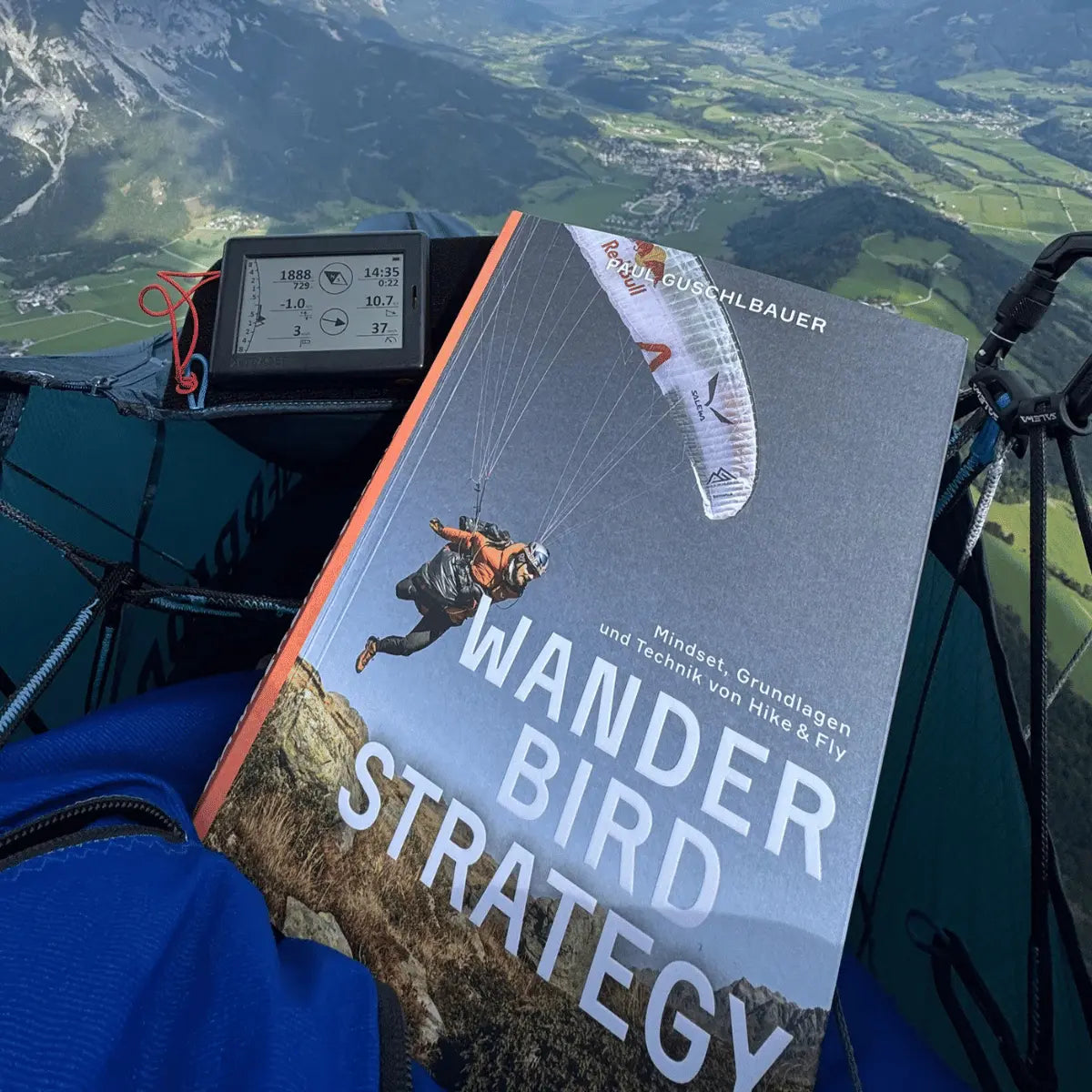 Buch "Wanderbird Strategie von Paul Guschlbauer" liegt im Gleitschirm-Hike-&-Fly-Equipment, neben einem Variometer mit Bergpanorama im Hintergrund.