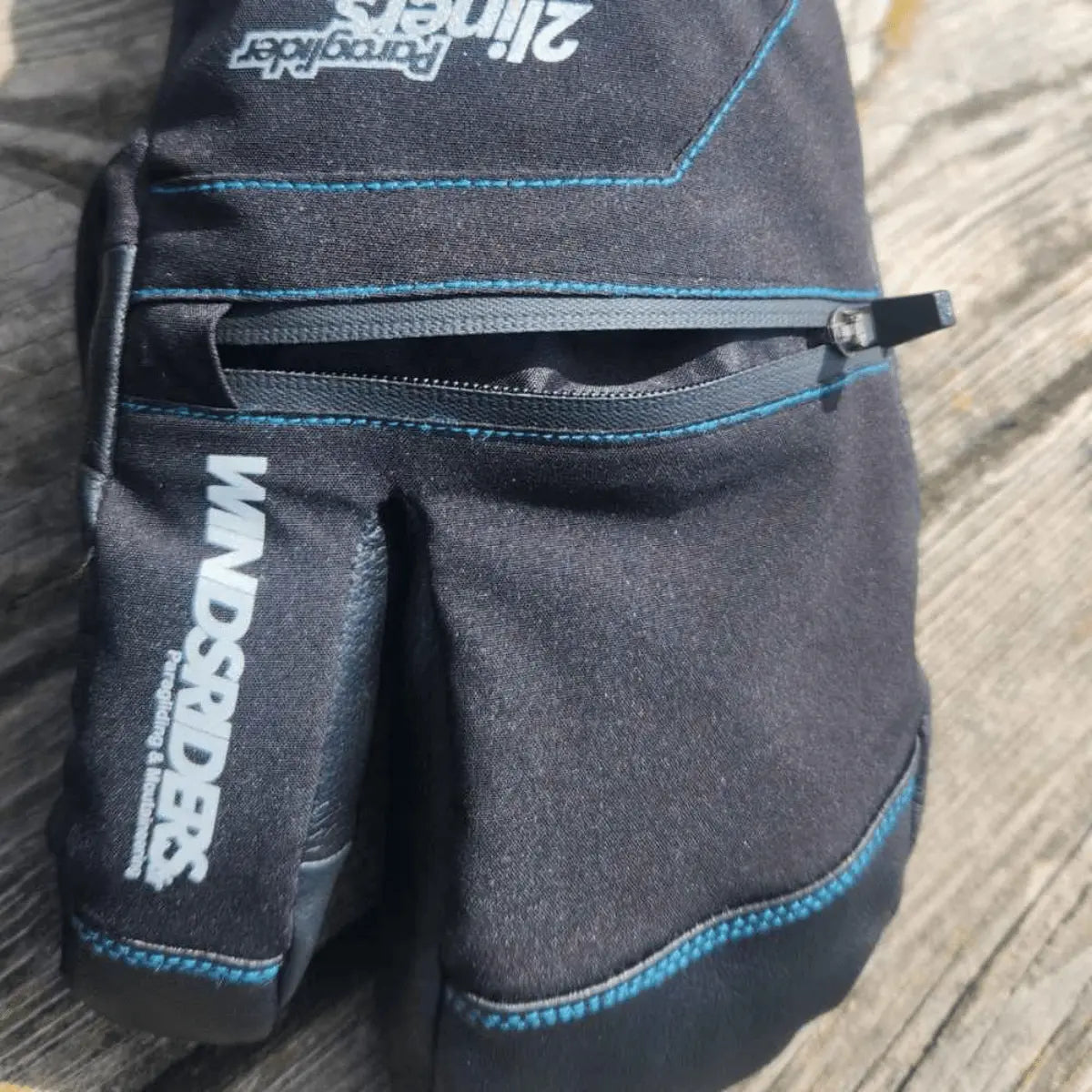 Schwarzer Handschuh mit drei offenen Fingern und blauer Stickerei auf einem Holztisch, entwickelt für Gleitschirmpiloten.