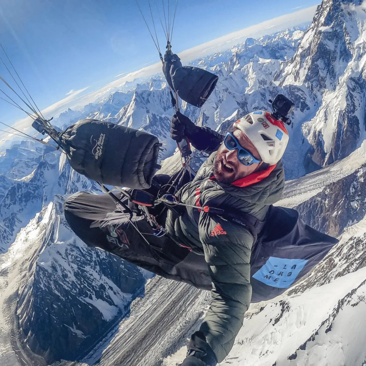 Paraglider in action high above snow-capped mountains, wearing a helmet and goggles, with a GoPro camera attached.