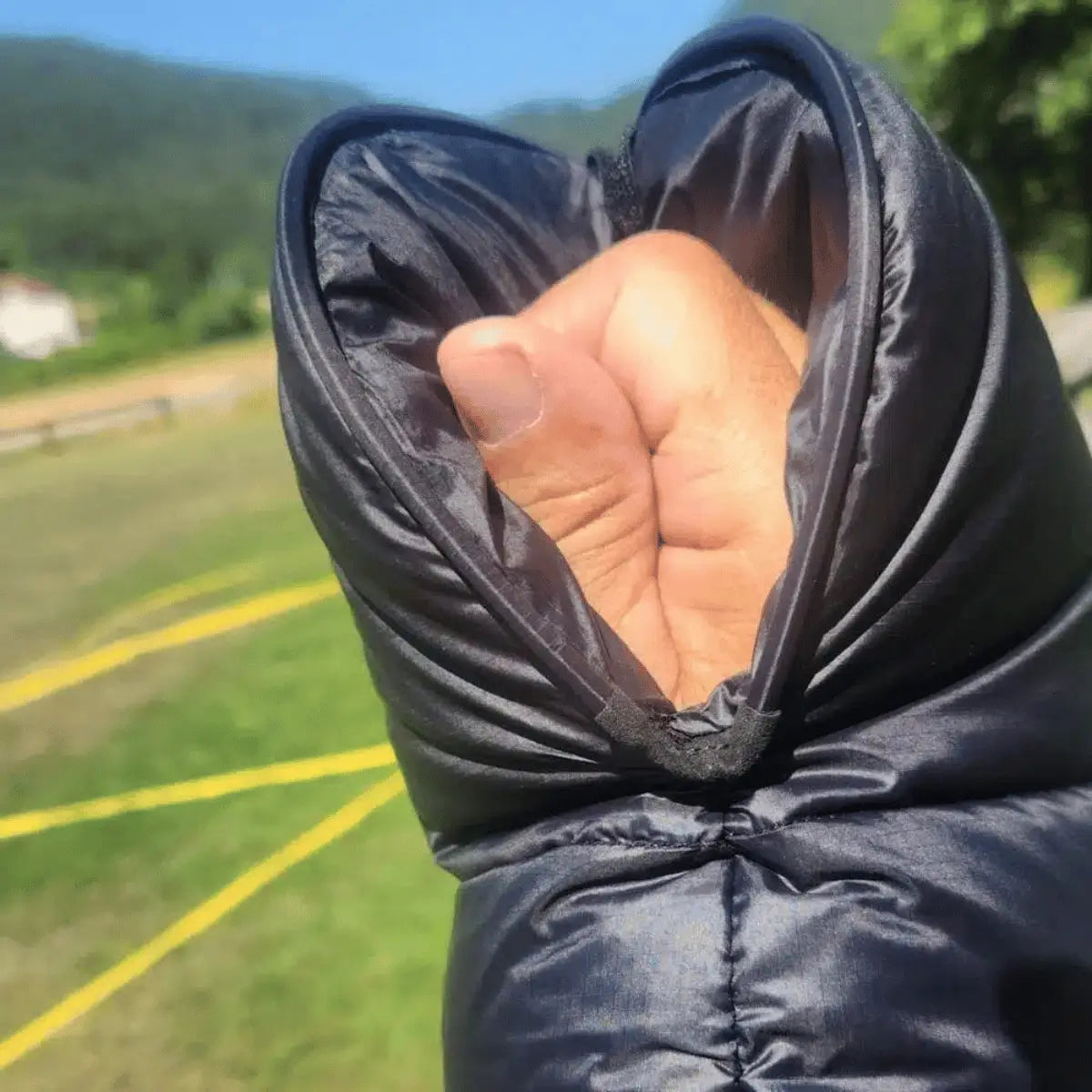 Windsriders K2 Magnet Muffles: Schwarze, wattierte Ärmel mit Magnetverschluss, die eine Hand wärmen, mit Blick auf eine grüne Hügellandschaft.