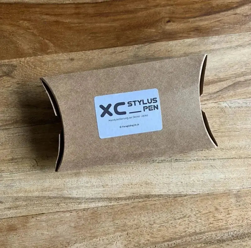 XC StylusPen - Perfekt für die Jacke! Ein brauner Karton mit blauem Etikett auf Holz, enthält den praktischen StylusPen für einfaches Handy-Bedienen beim Gleitschirmfliegen.