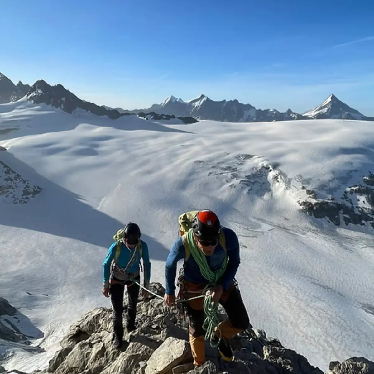 Zwei Bergsteiger steigen auf einem felsigen Grat mit Seilen gesichert, im Hintergrund eine weite Gletscherlandschaft unter blauem Himmel.