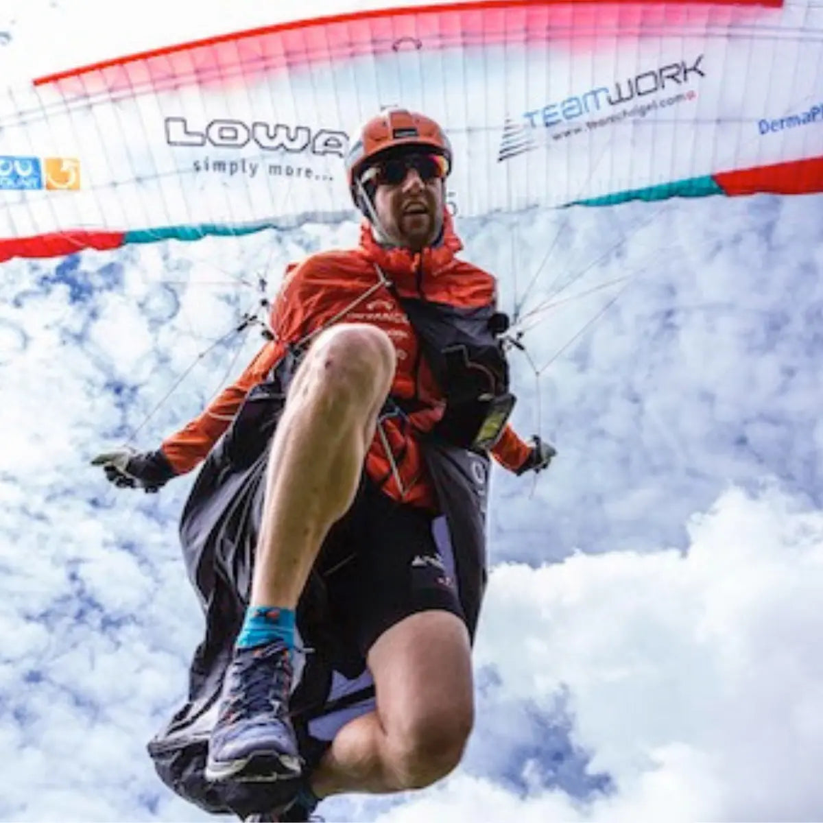 Mann im orangefarbenen Gleitschirm-Outfit fliegt mit seinem Gleitschirm "XPeaks - Neue Wege auf alte Berge!" vor blauem Himmel.