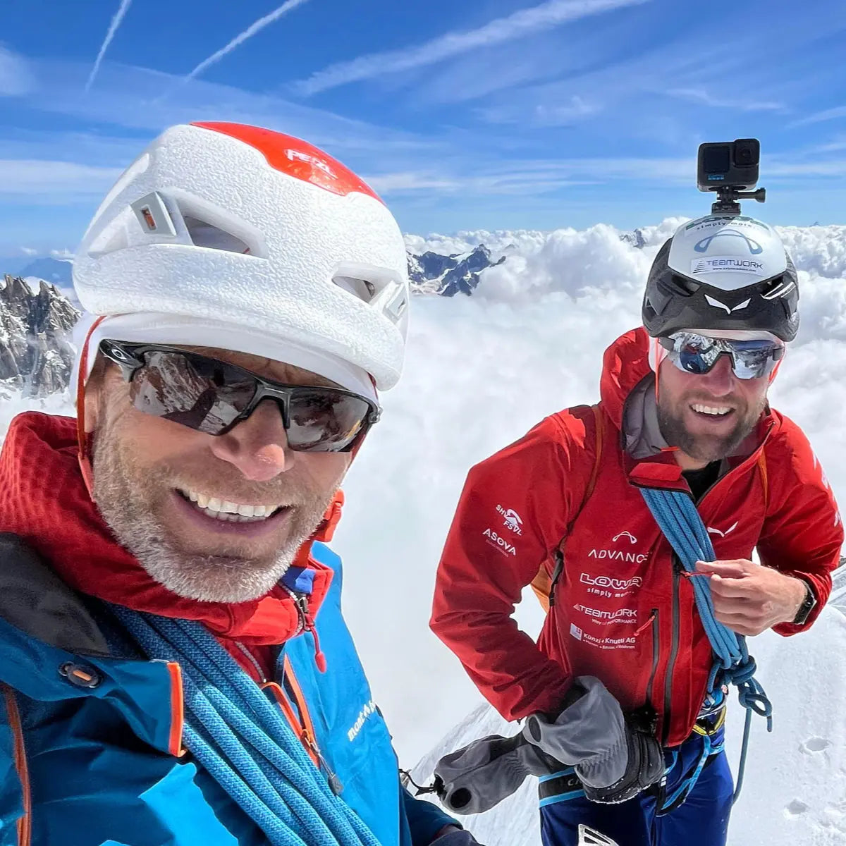 Zwei Bergsteiger mit XPeaks Helmen und Ausrüstung auf einem schneebedeckten Gipfel mit Wolken im Hintergrund.