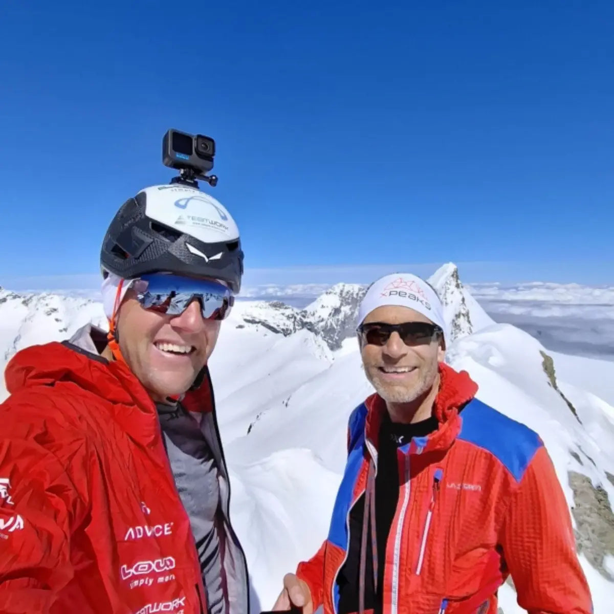 Zwei Männer in roten Jacken und mit Helmen auf einem verschneiten Berggipfel, bereit für neue Wege auf alte Berge mit XPeaks.