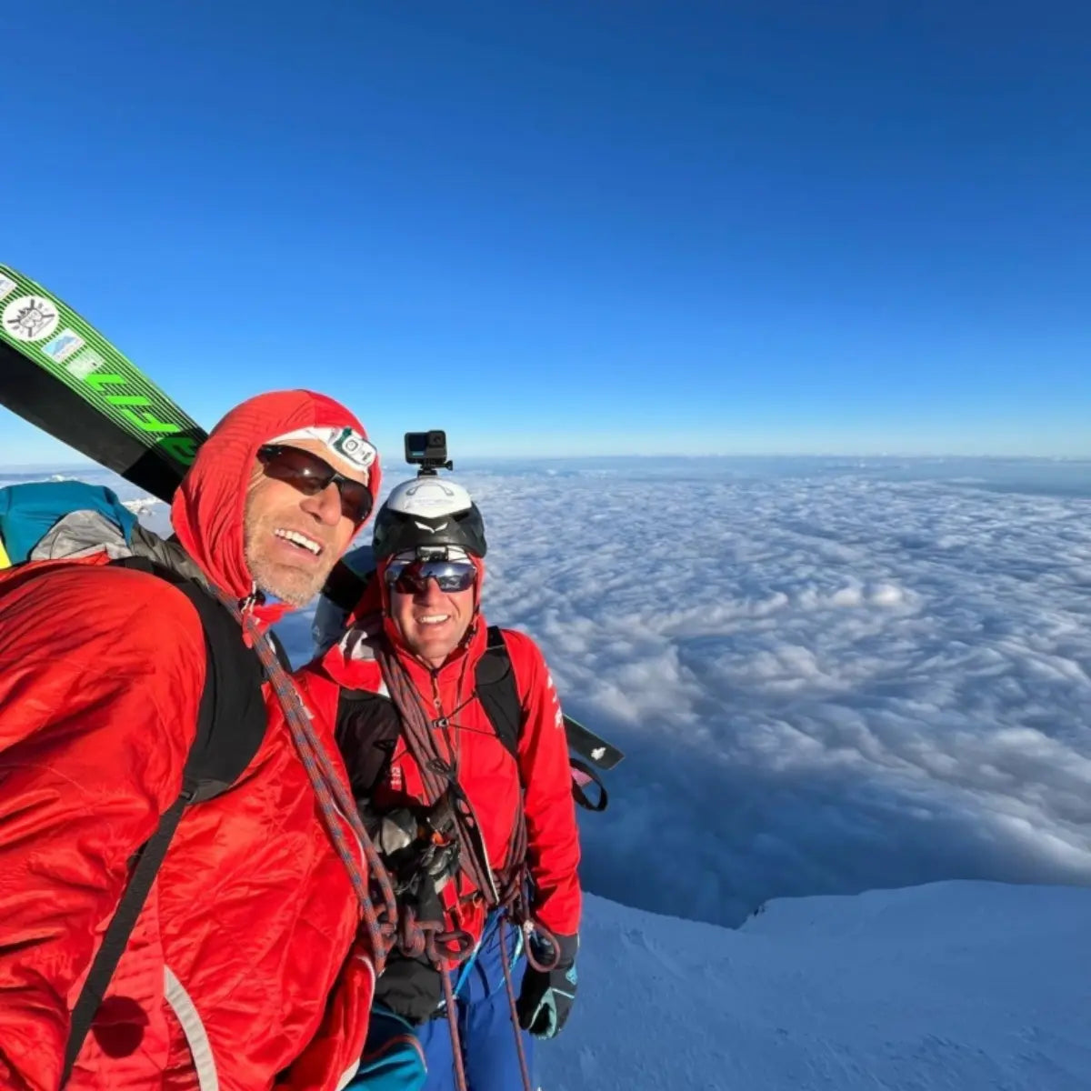 Zwei Bergsteiger mit Skiern und Ausrüstung auf einem schneebedeckten Gipfel über einer Wolkendecke, bereit für neue Wege mit XPeaks.