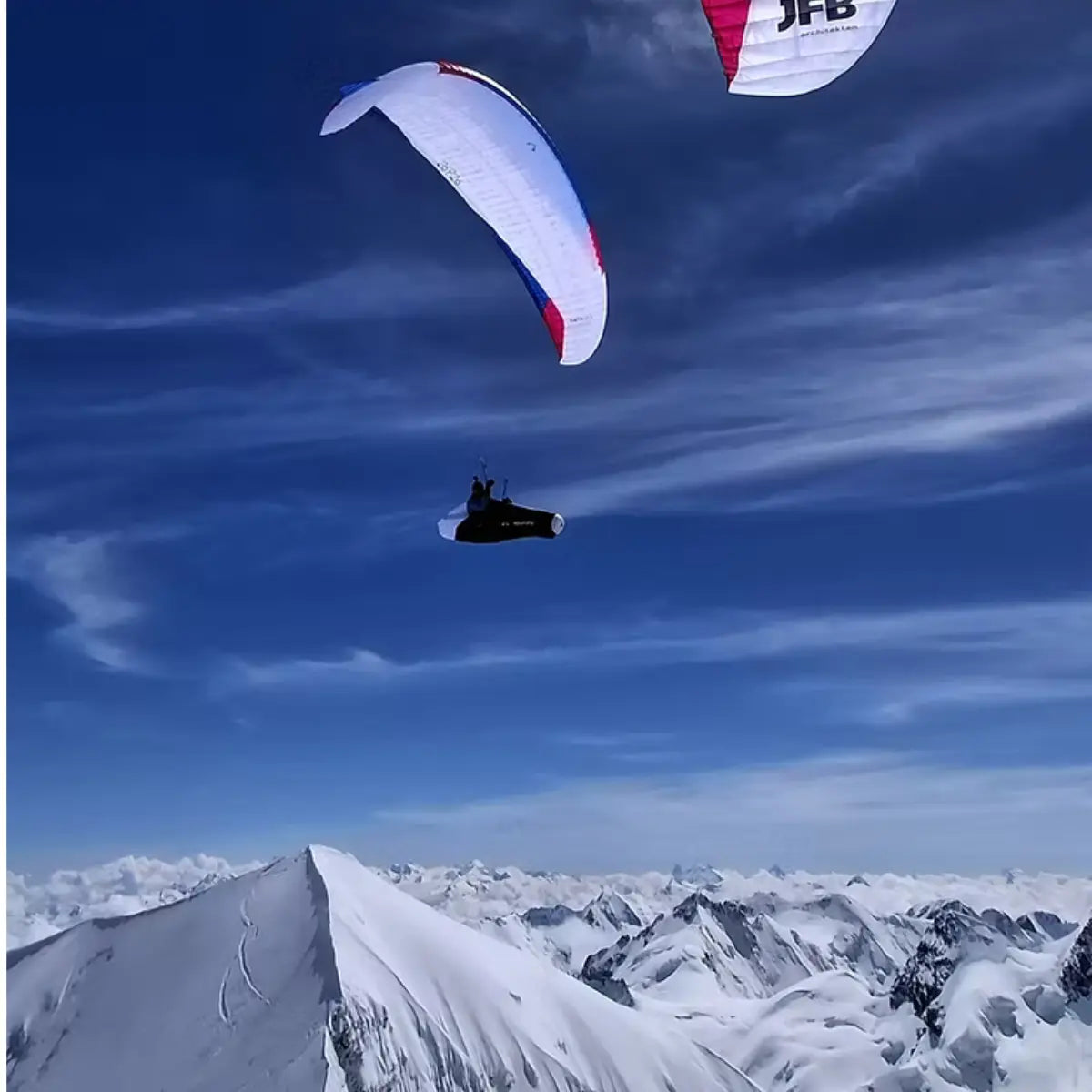 Paraglider pilot soaring over snow-capped mountains, with a red and white canopy visible in the sky. The XPeaks paraglider offers new ways to explore old mountains.